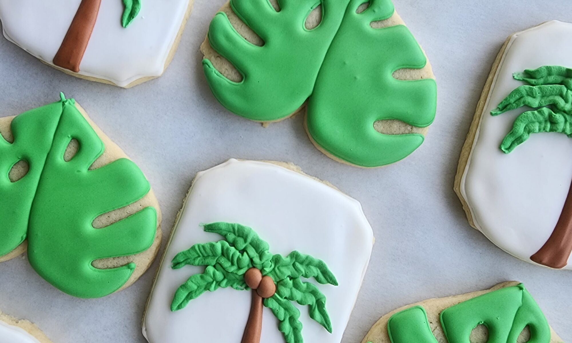 Decorated sugar cookies shaped like palm trees and tropical monstera leaves, arranged on a baking sheet.