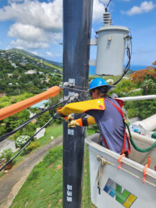 Lineworker in a blue hard hat and insulated rubber sleeves working on power lines from a bucket truck, with a transformer overhead and green hillside homes and the Caribbean Sea behind him.