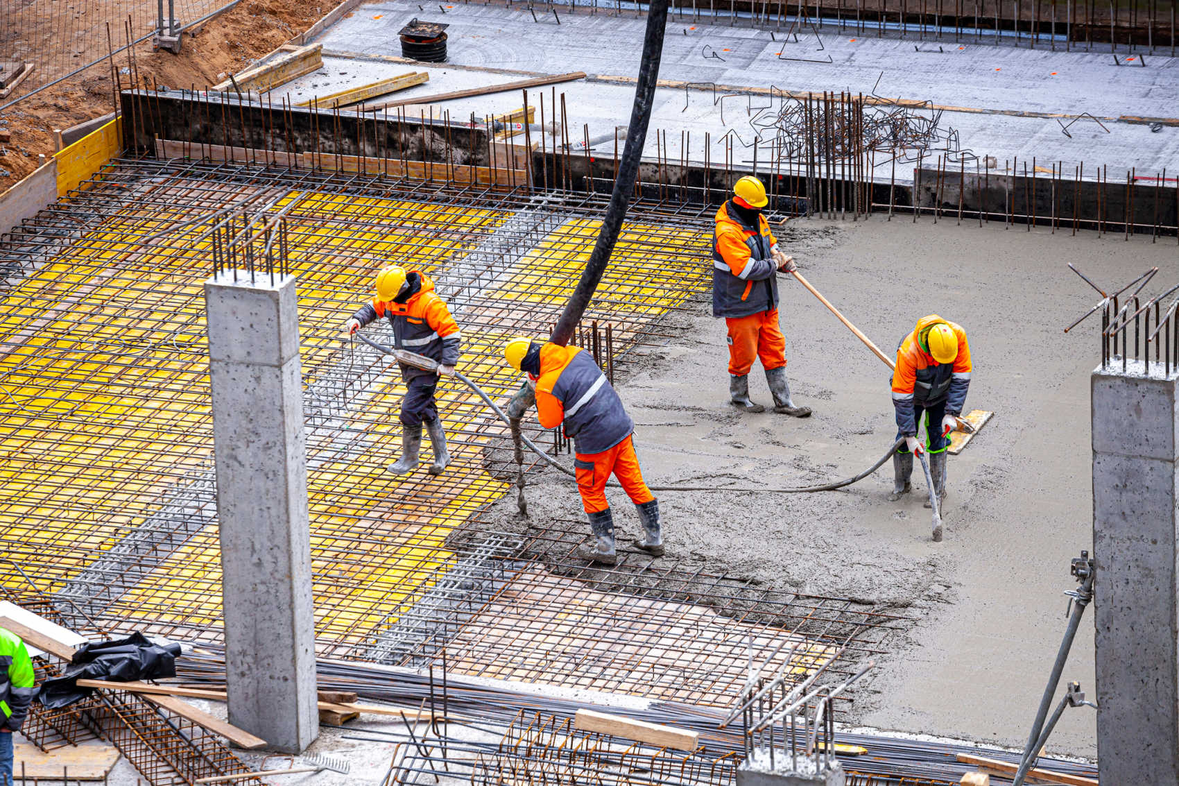 Four construction workers in orange safety gear and yellow hard hats pouring and leveling concrete over a steel rebar grid on a building foundation.