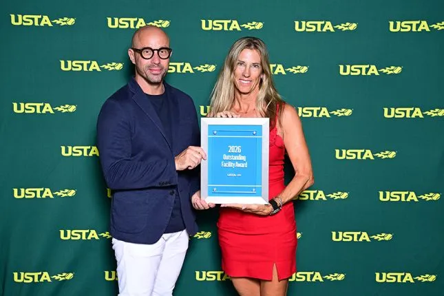 Dena Coe, Director of Tennis at The Buccaneer, in a red dress, holding a blue plaque reading "2026 Outstanding Facility Award" with a man in a navy blazer in front of a green USTA backdrop.