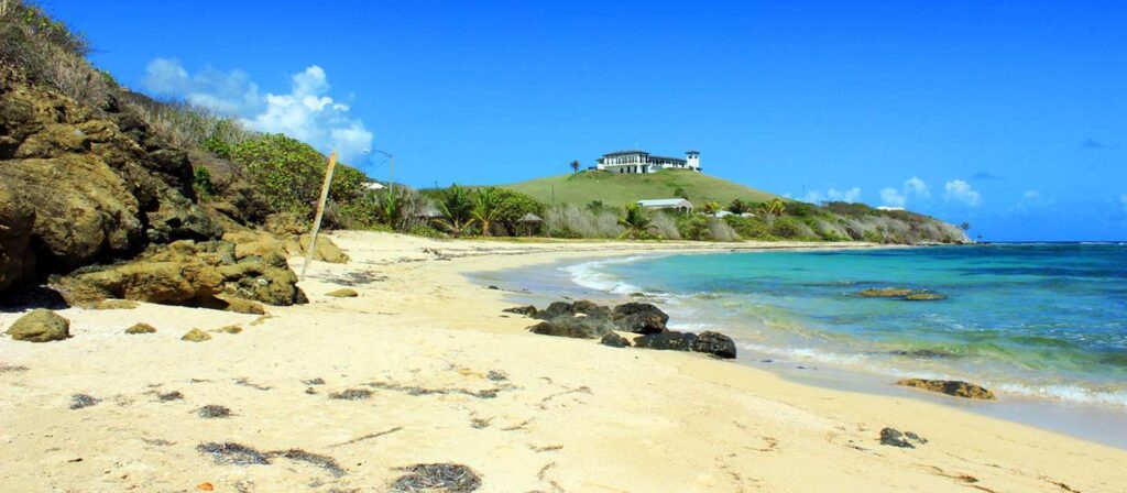 Sandy beach with rocky outcrops and turquoise water on St. Croix, with a large white house on a green hilltop in the distance under a blue sky.